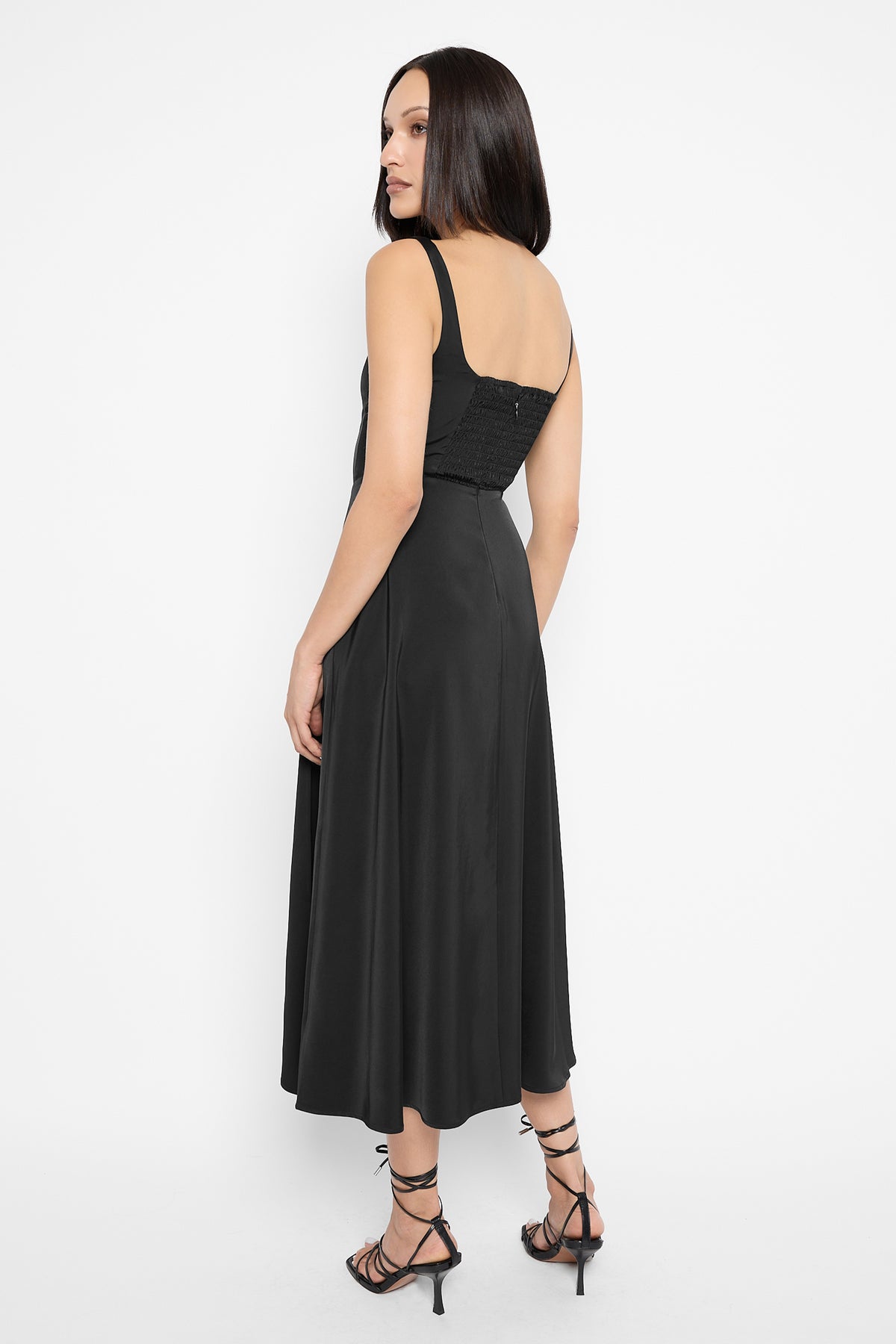 Back view of a model looking over her shoulder, wearing the black Alyx satin midi dress with a smocked back panel, a flowing skirt and black strappy lace-up stiletto sandals.