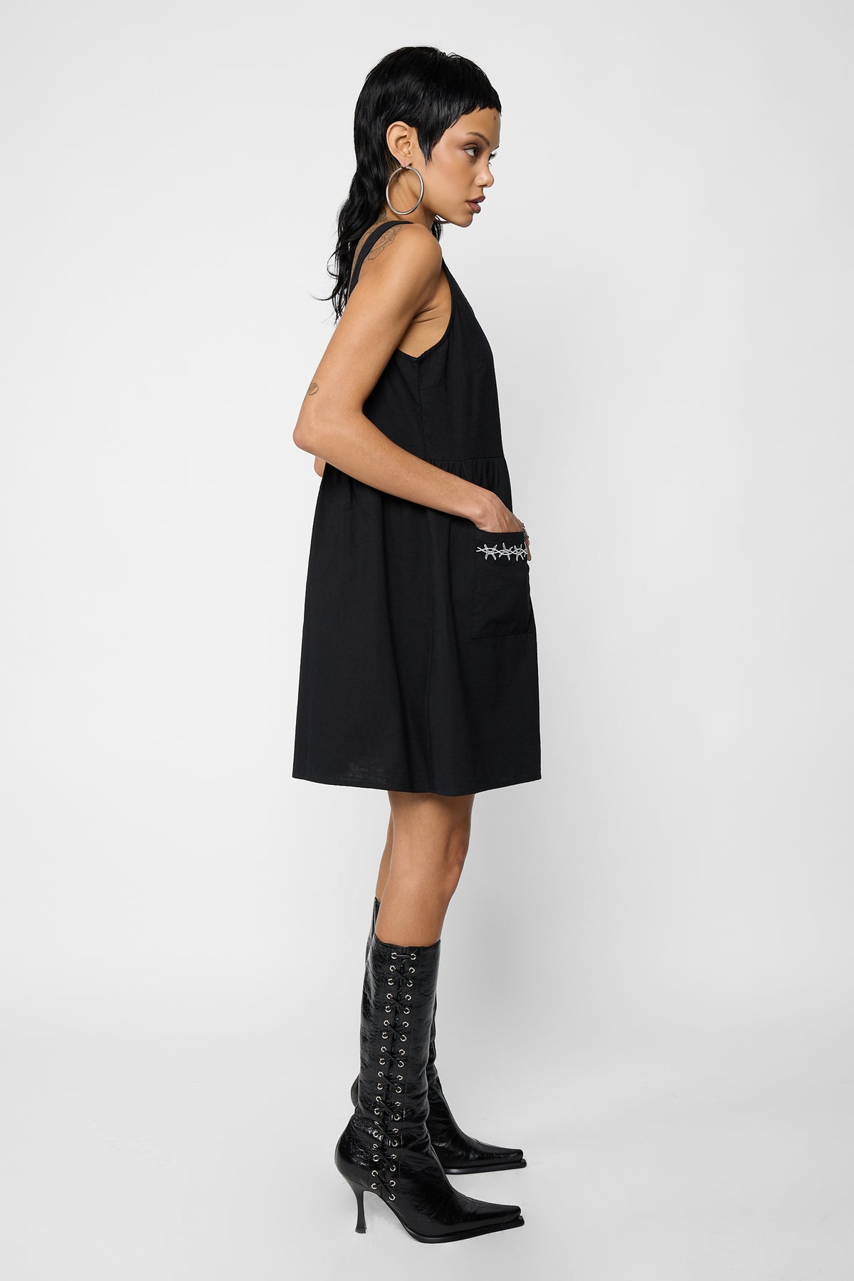 Side view of a model with a hand in the barbed-wire-embroidered patch pocket of the black Cora smock dress, wearing black lace-up pointed knee-high boots.