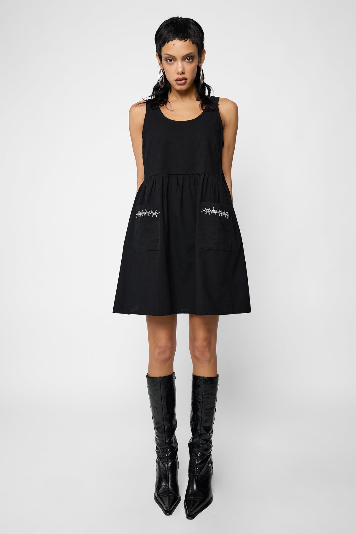 Full-length front view of a model wearing the black sleeveless Cora smock dress with white barbed-wire embroidery on the patch pockets and black pointed knee-high boots.