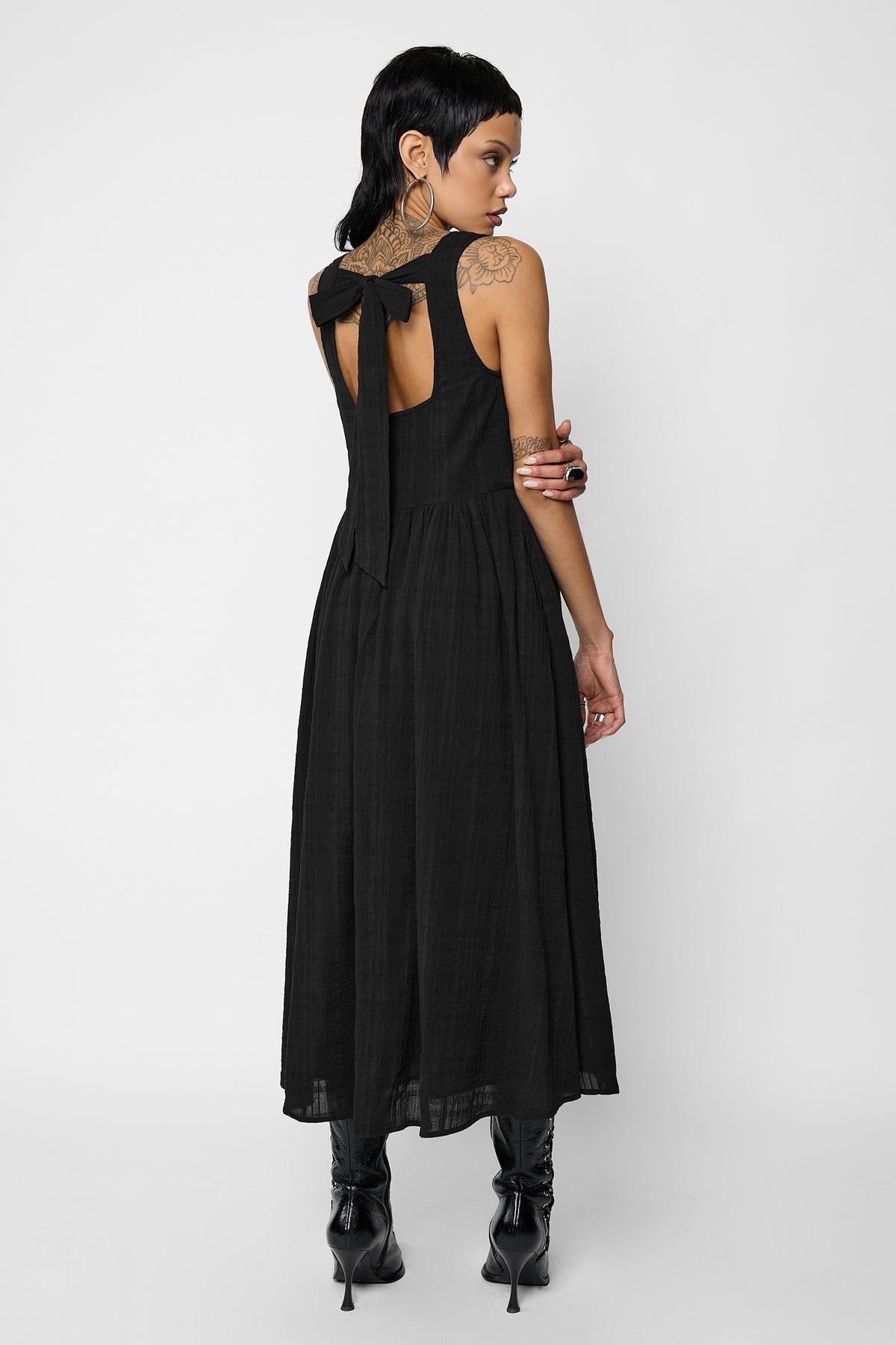 Back view of a model looking over her shoulder, wearing the black Esme smock midi dress with an open back tied with a bow and a gathered midi skirt, with black stiletto ankle boots.