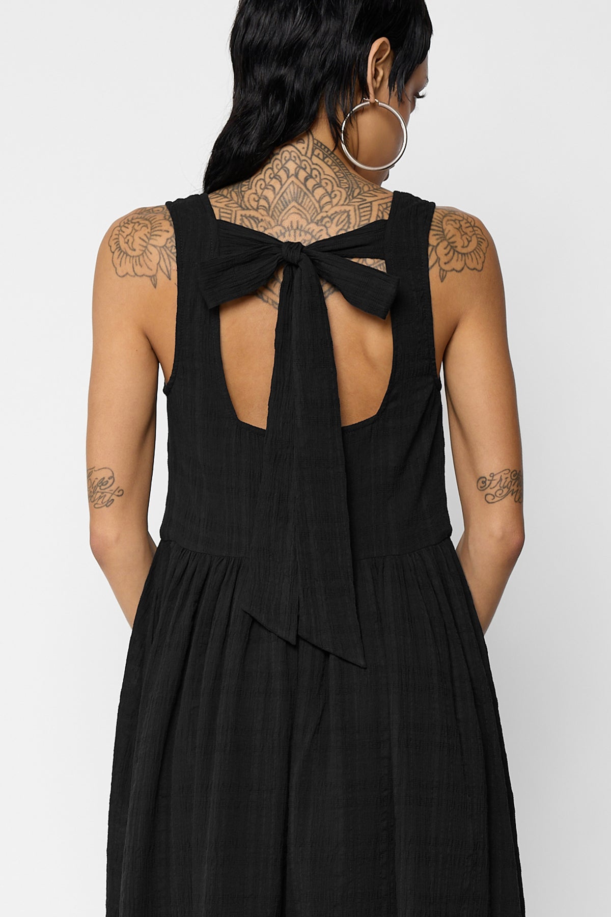 Close-up of the open back of the black Esme dress with a bow tied at the top and long ribbon ties falling down the back, with the gathered skirt below.