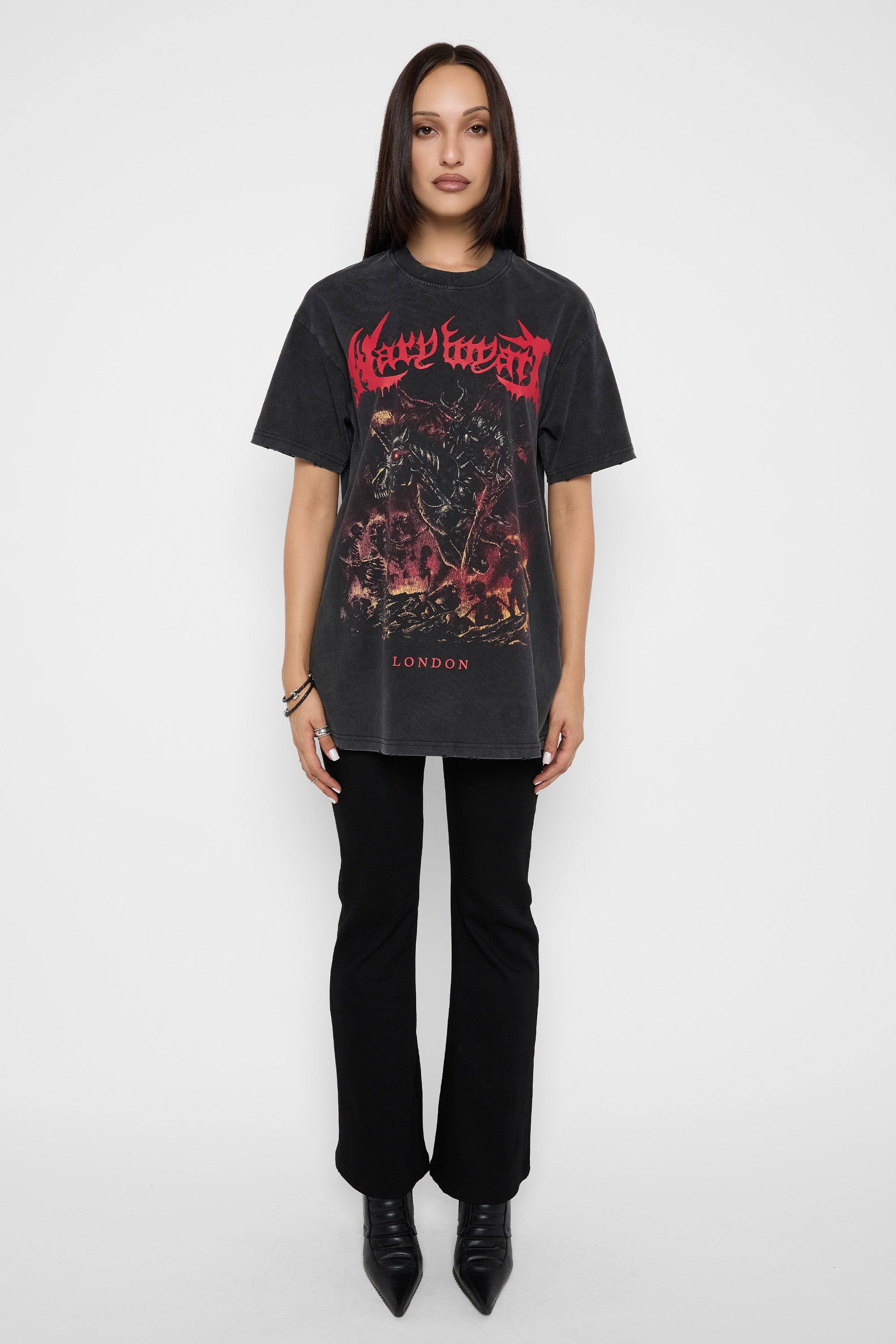 A model stands against a white studio background wearing a black graphic t-shirt with a red and black 'Mary Wyatt' print, paired with black pants and black pointed boots.