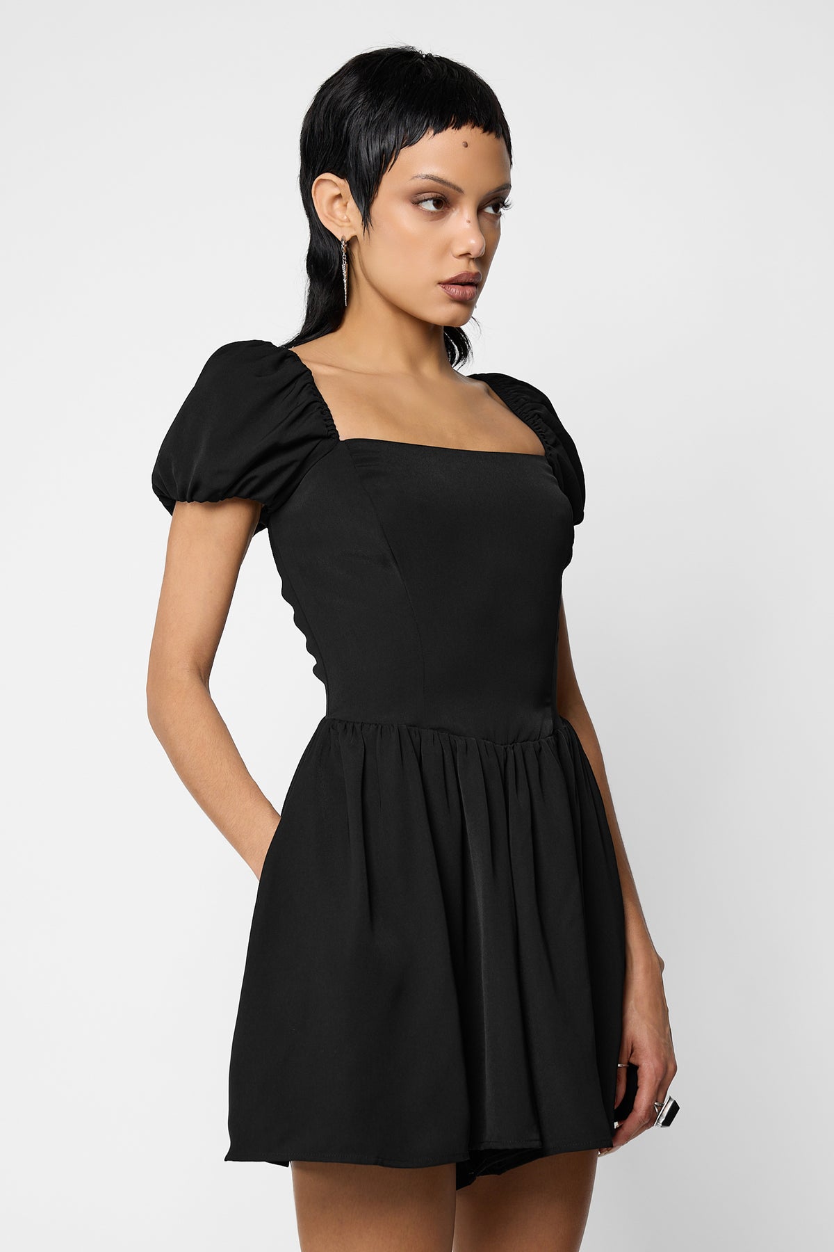 Three-quarter view of a model looking aside, wearing the black Nevermore playsuit with a square neckline, puff sleeves, a fitted bodice and a gathered skirt-style short.