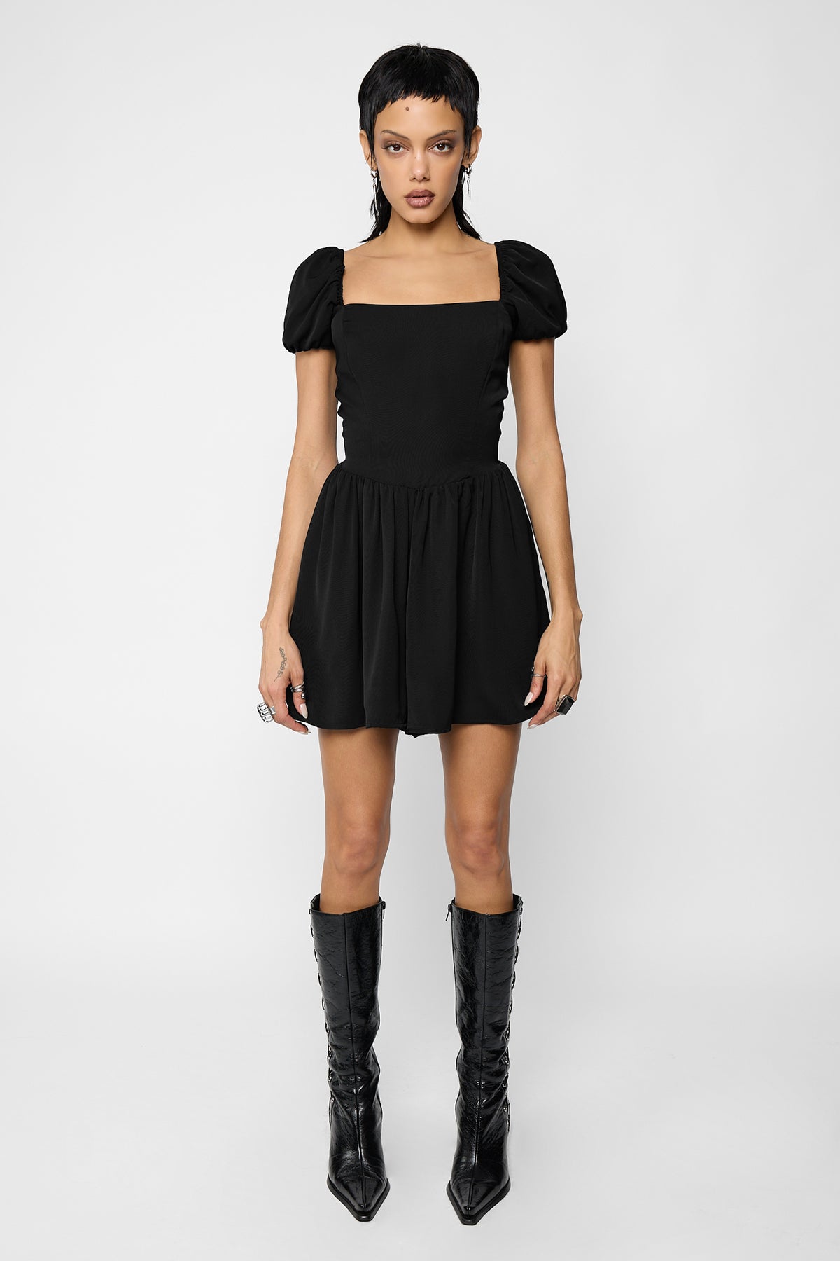 Full-length front view of a model with a short black mullet wearing the Nevermore lace-up playsuit, a black puff-sleeve square-neck playsuit with a fitted bodice and a gathered skirt-style short, with black pointed lace-up knee-high boots.