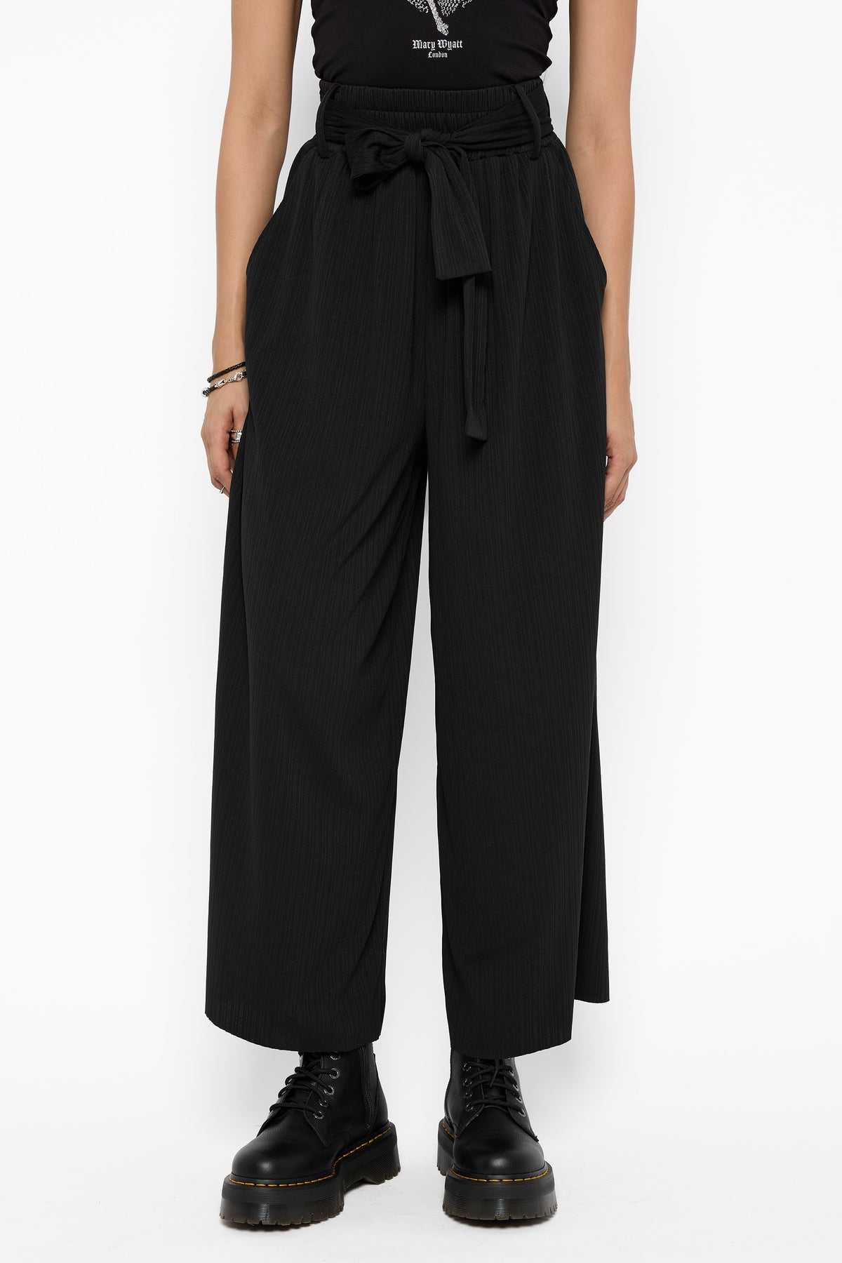 Front view from the chest down of the black Rowan wide-leg culottes with a tied sash at the high waist, side pockets and black platform lace-up boots.