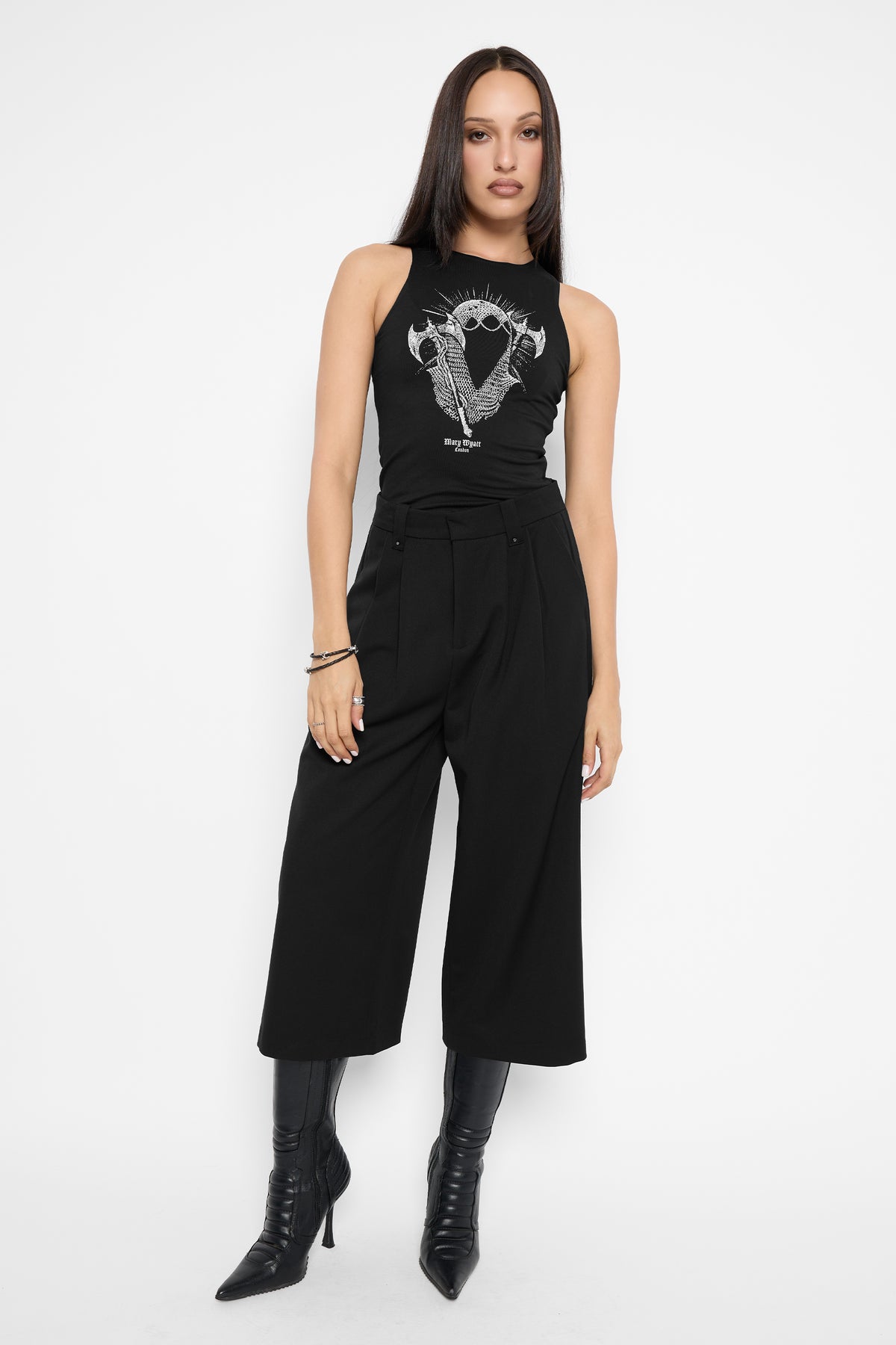 Full-length front view of a model with long dark hair wearing the black Tegan cropped wide-leg trousers with a black racer tank with a white chainmail-hooded figure between two axes, and pointed black knee-high boots.
