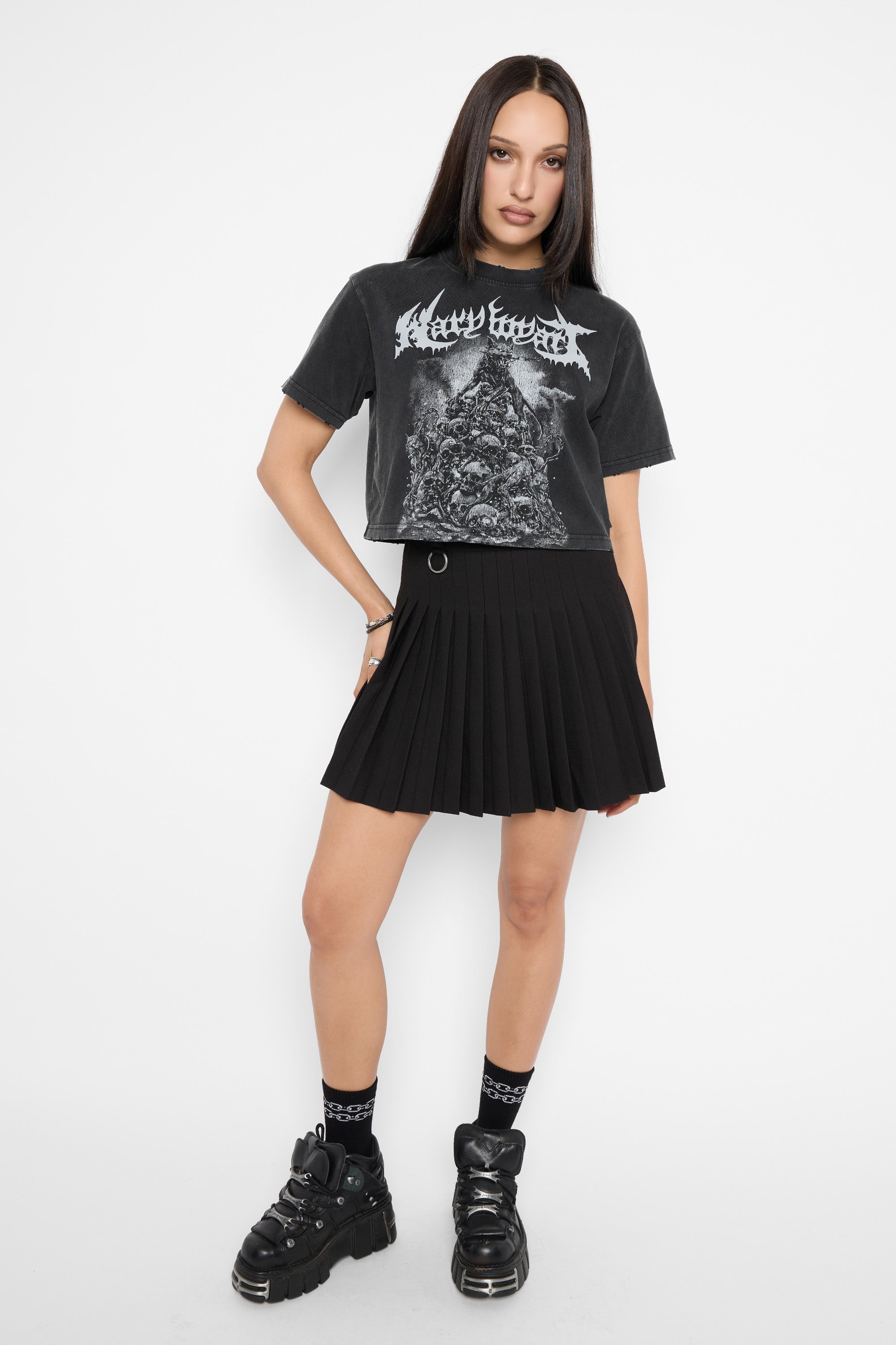 A model wearing a black short-sleeve graphic t-shirt with a white metal logo, a black pleated mini skirt, black socks, and black platform boots, standing against a plain white background.