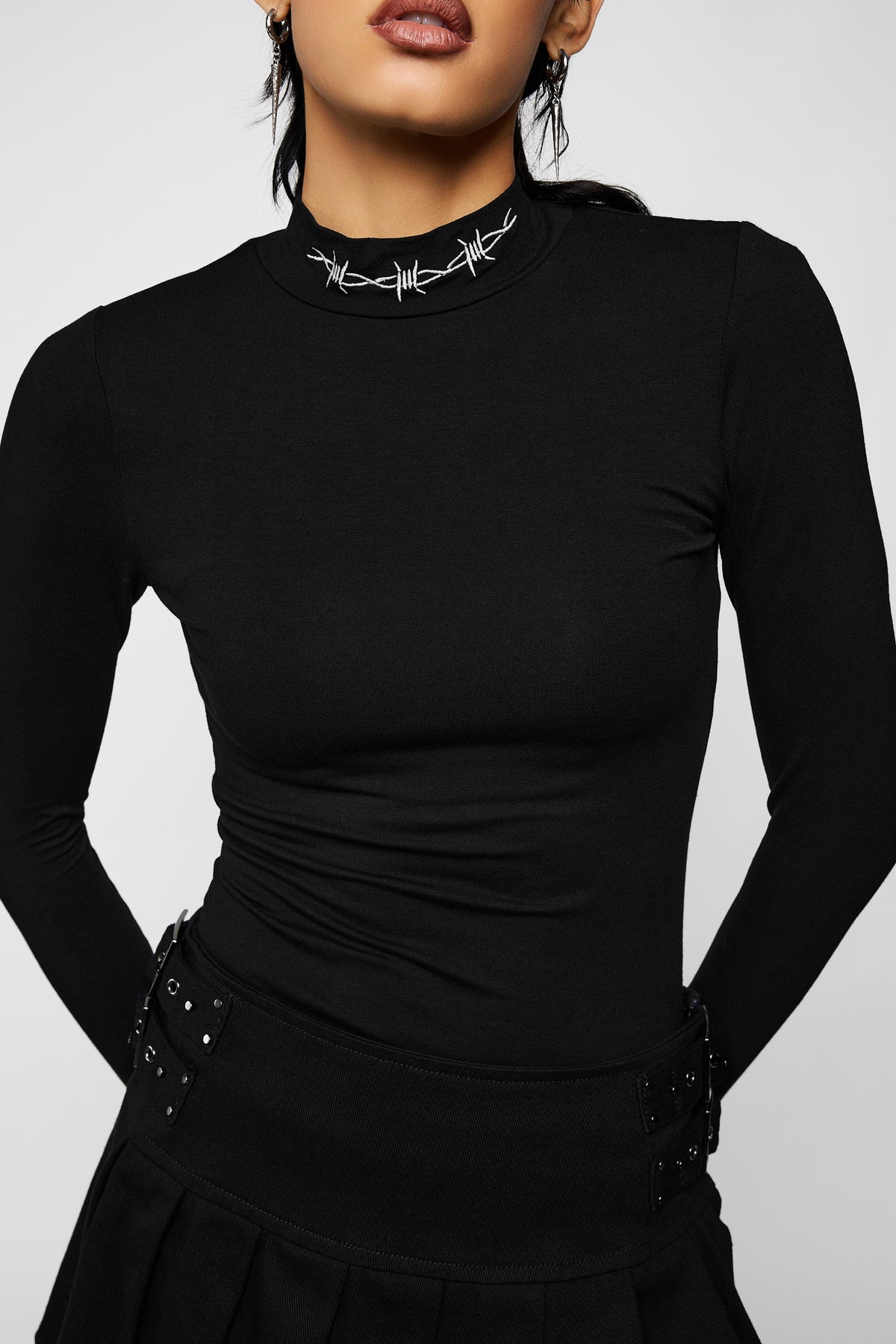 Close-up of the black Bound long sleeve top's white embroidered barbed-wire collar, with the button waistband and D-ring of the black trousers.