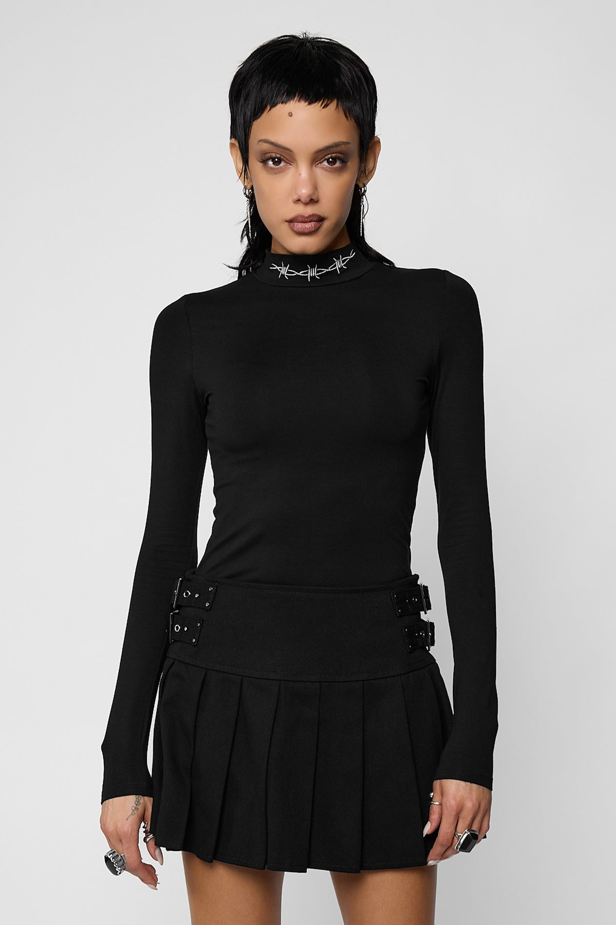 A model with long dark hair wearing the black Bound high neck long sleeve III, a fitted mock-neck top with a white embroidered barbed-wire collar, with black high-waisted trousers with a button waist and a D-ring.