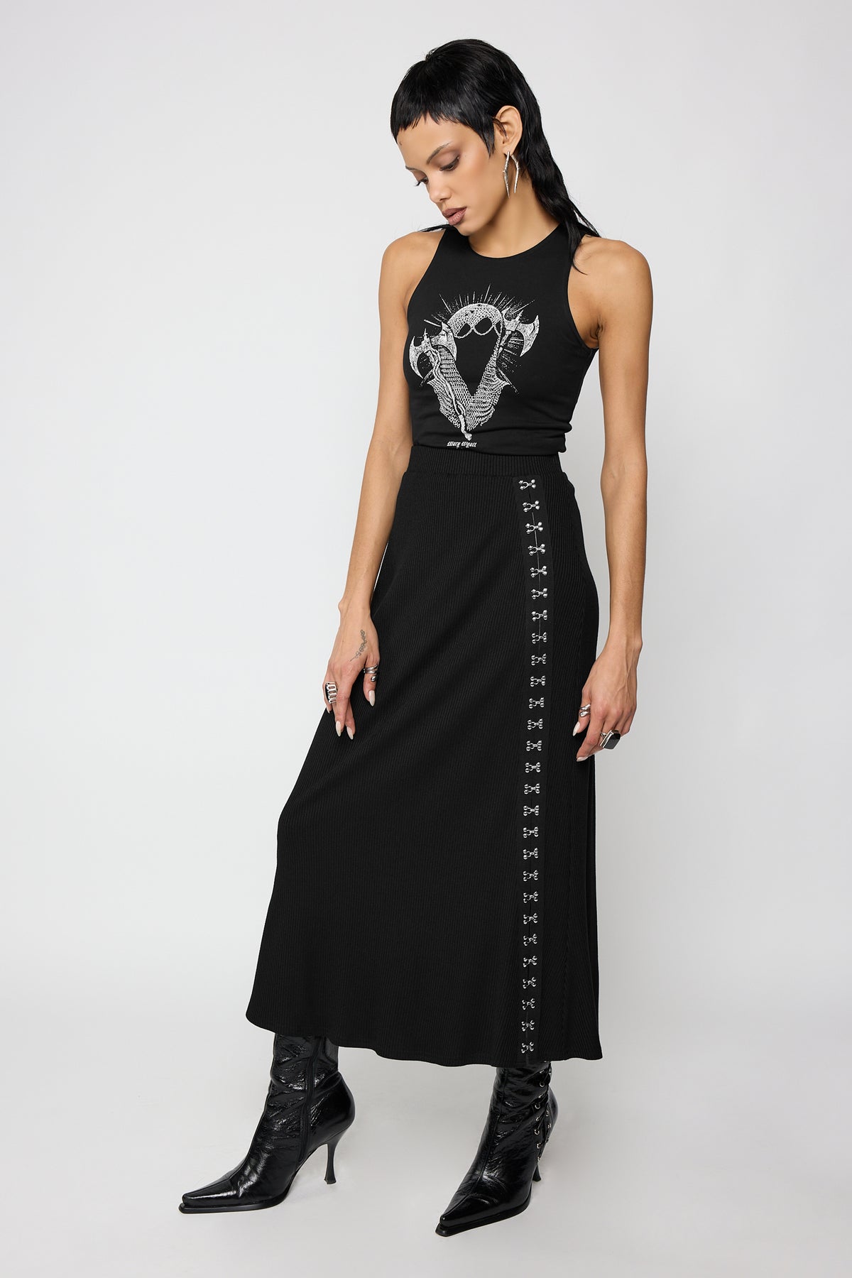 A model wearing a black sleeveless with a white graphic print on the chest, styled with the beatriz skirt, a black ribbed skirt with hook and eye fastenings from hip to ankle, photographed against a light gray studio background.