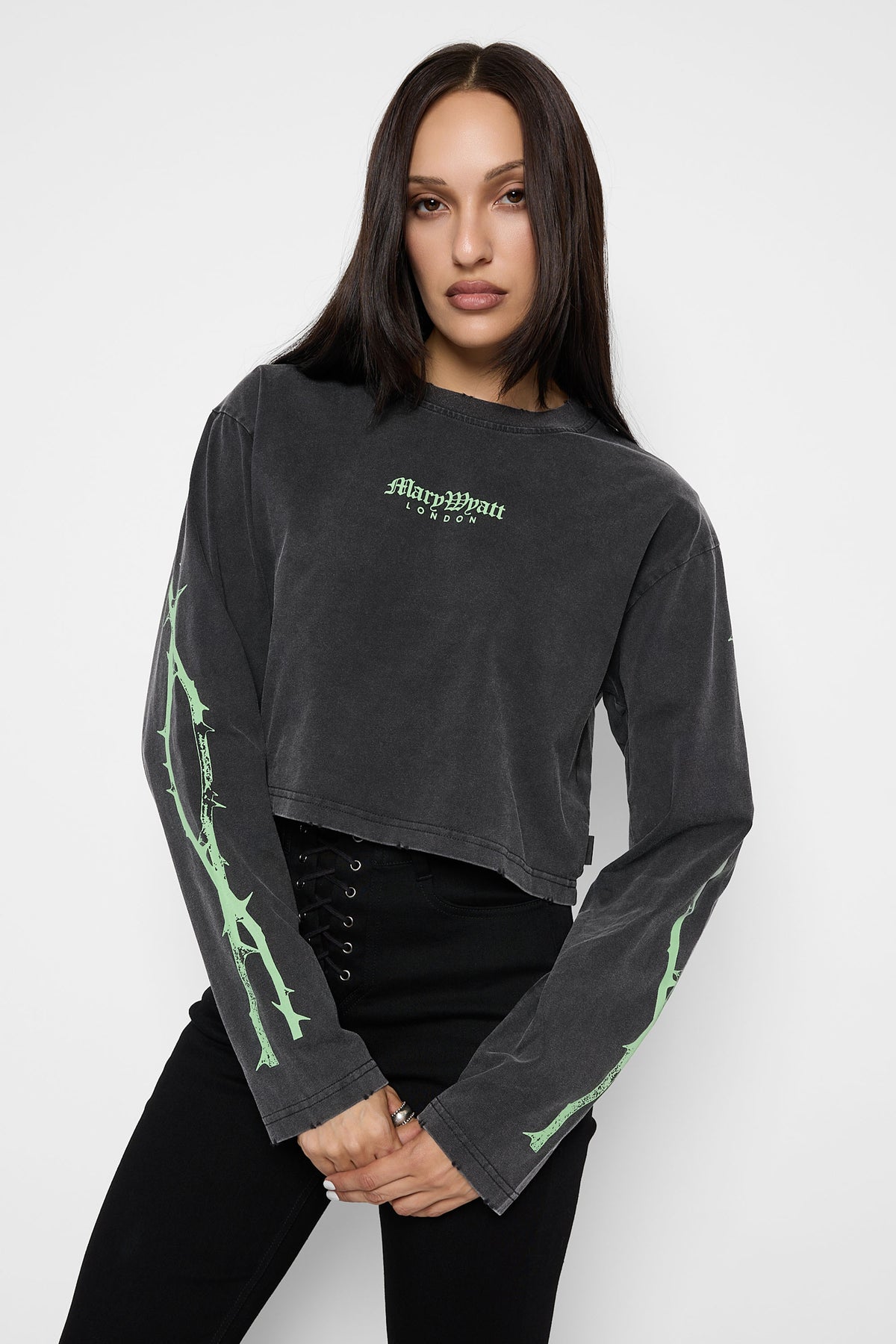 A model with her hands clasped wearing the washed charcoal Druidess cropped long sleeve tee with pale green "Mary Wyatt London" lettering and thorny sleeve prints, with black lace-up trousers.