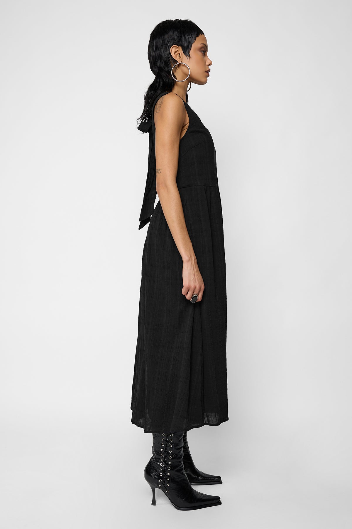 Side profile of a model wearing the black Esme smock midi dress with the back bow ribbon trailing down, with a large hoop earring and black lace-up stiletto ankle boots.
