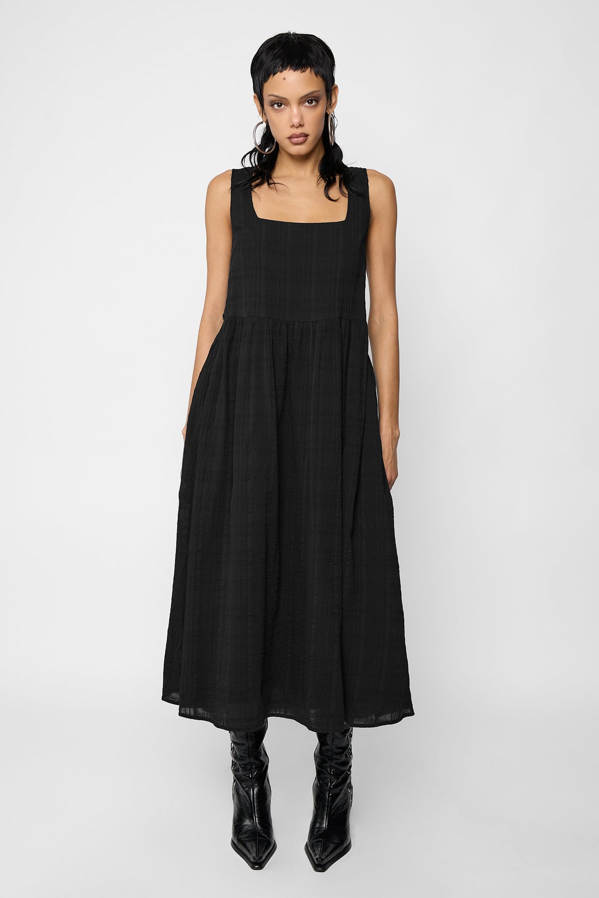 Full-length front view of a model with a short black mullet and hoop earrings wearing the Esme smock midi dress, a black sleeveless square-neck dress in a tonal checked fabric with a gathered skirt, with black pointed ankle boots.