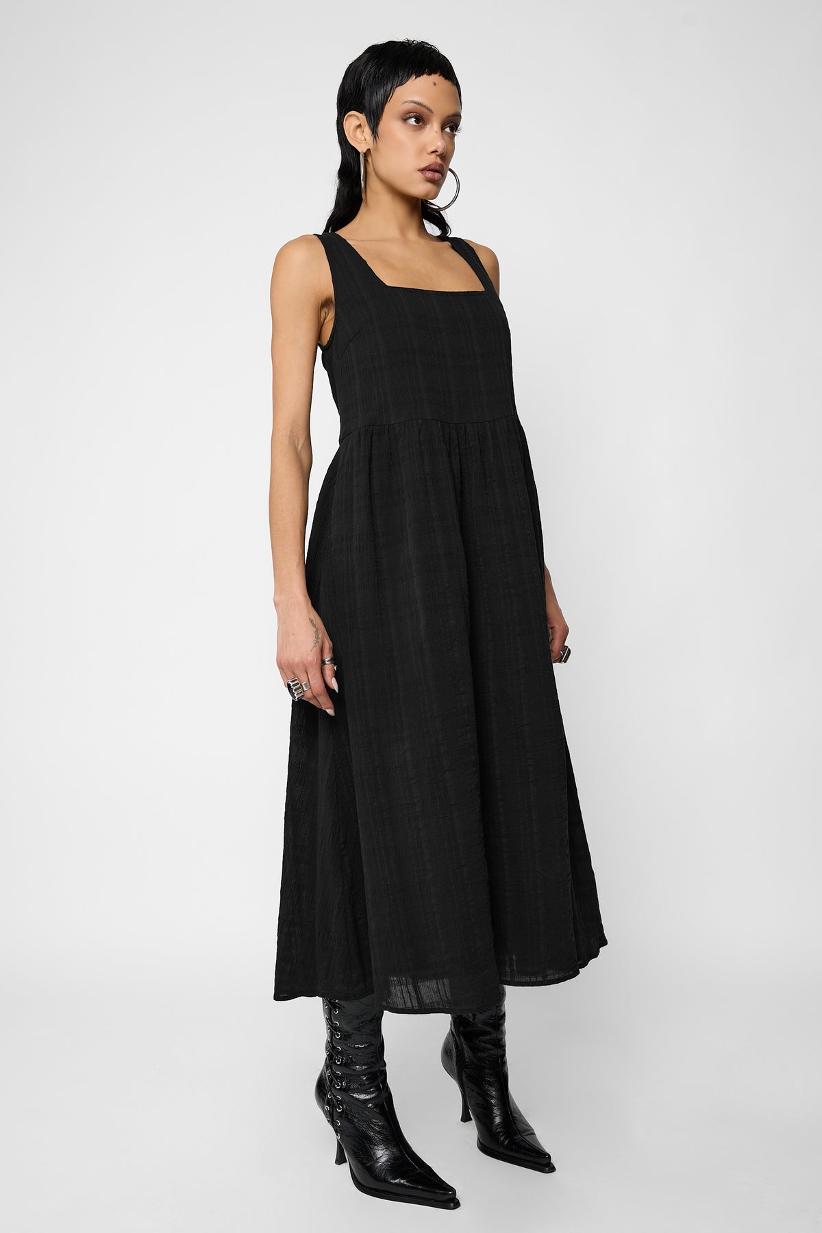 Three-quarter front view of a model wearing the black sleeveless Esme smock midi dress with a square neckline and gathered skirt, with pointed black lace-up ankle boots.