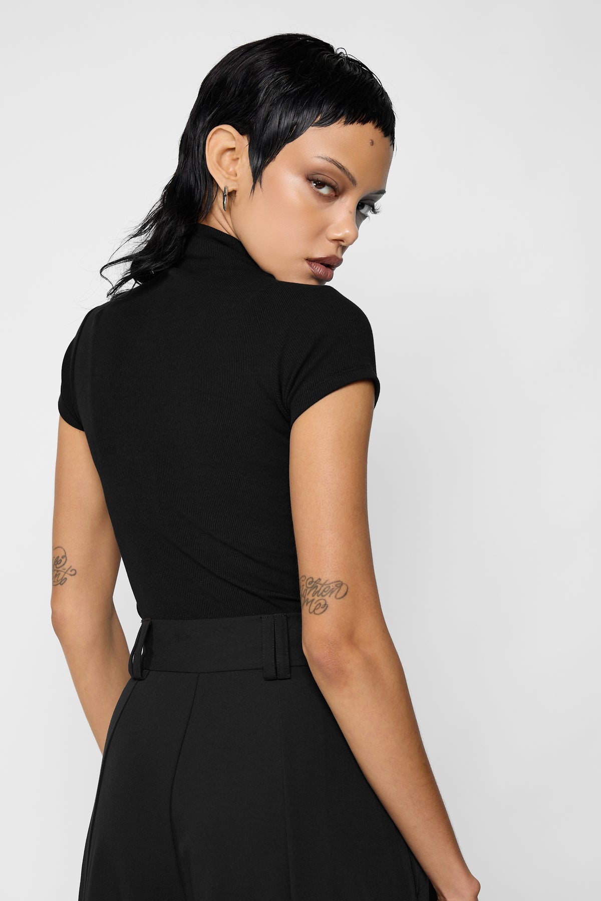 Back view of a model looking over her shoulder, wearing the plain black ribbed Madison top with cap sleeves and black high-waisted wide-leg trousers.