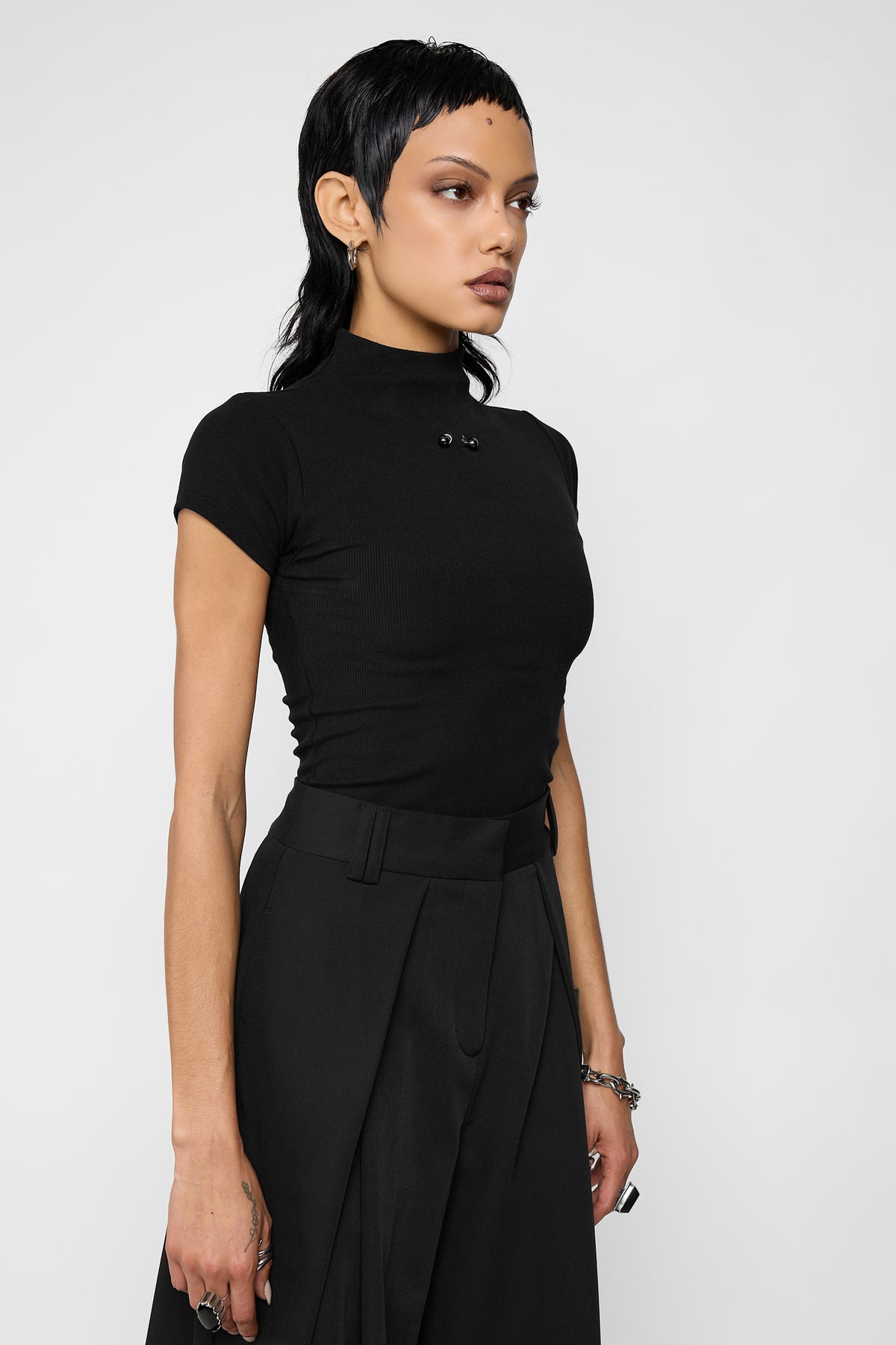 Three-quarter side view of a model wearing the black Madison mock-neck top with its ball barbell studs at the neckline, black pleated wide-leg trousers and a chain bracelet.