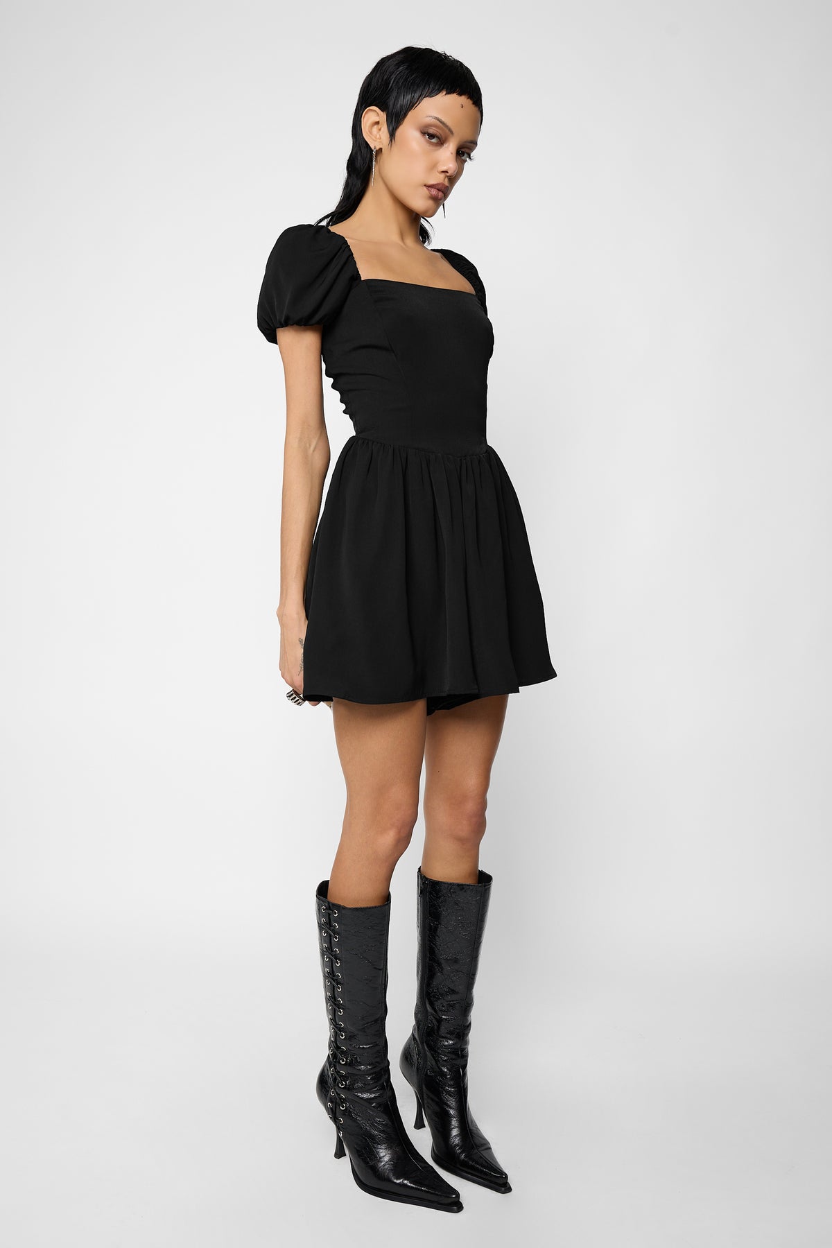 Three-quarter side view of a model wearing the black Nevermore playsuit with puff sleeves and black pointed lace-up knee-high boots.