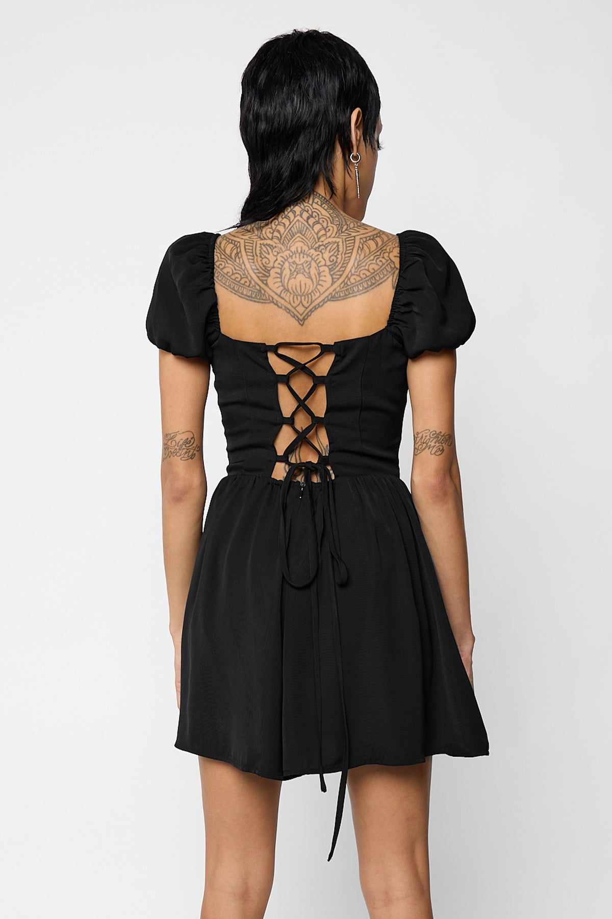 Back view of a model wearing the black Nevermore playsuit with an open lace-up back and long ties hanging down, with puff sleeves.