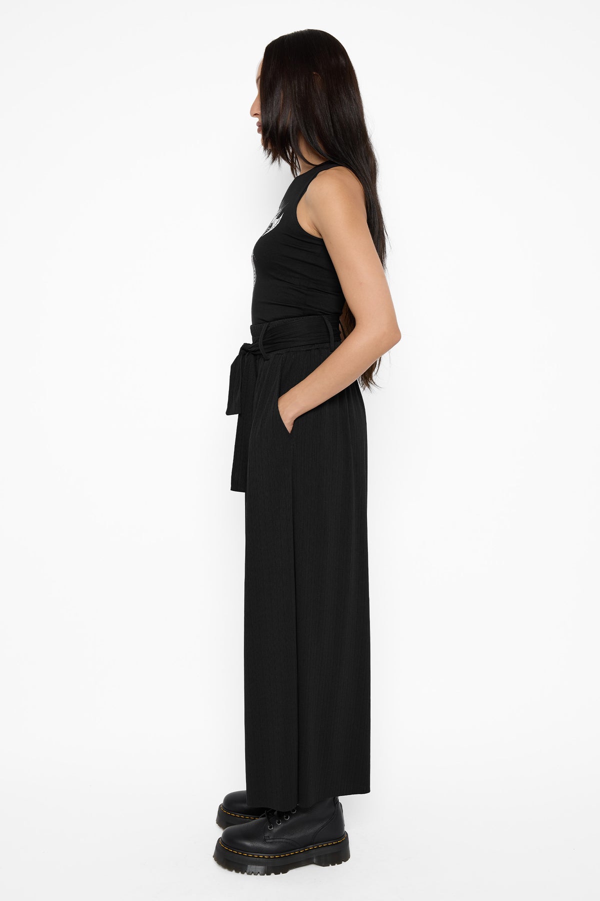 Side profile of a model with a hand in the pocket of the black Rowan culottes, with a tied sash at the waist, a black sleeveless top and black platform boots.