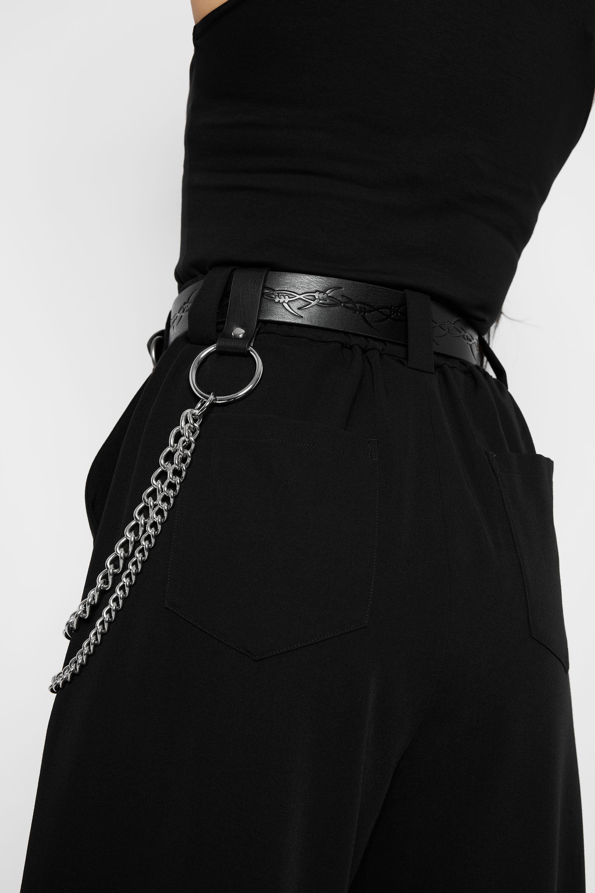 Close-up of a person wearing black wide-leg trousers and a black belt with a silver chain ring accessory hanging from the side, with embossed barbed wire on the belt leather.