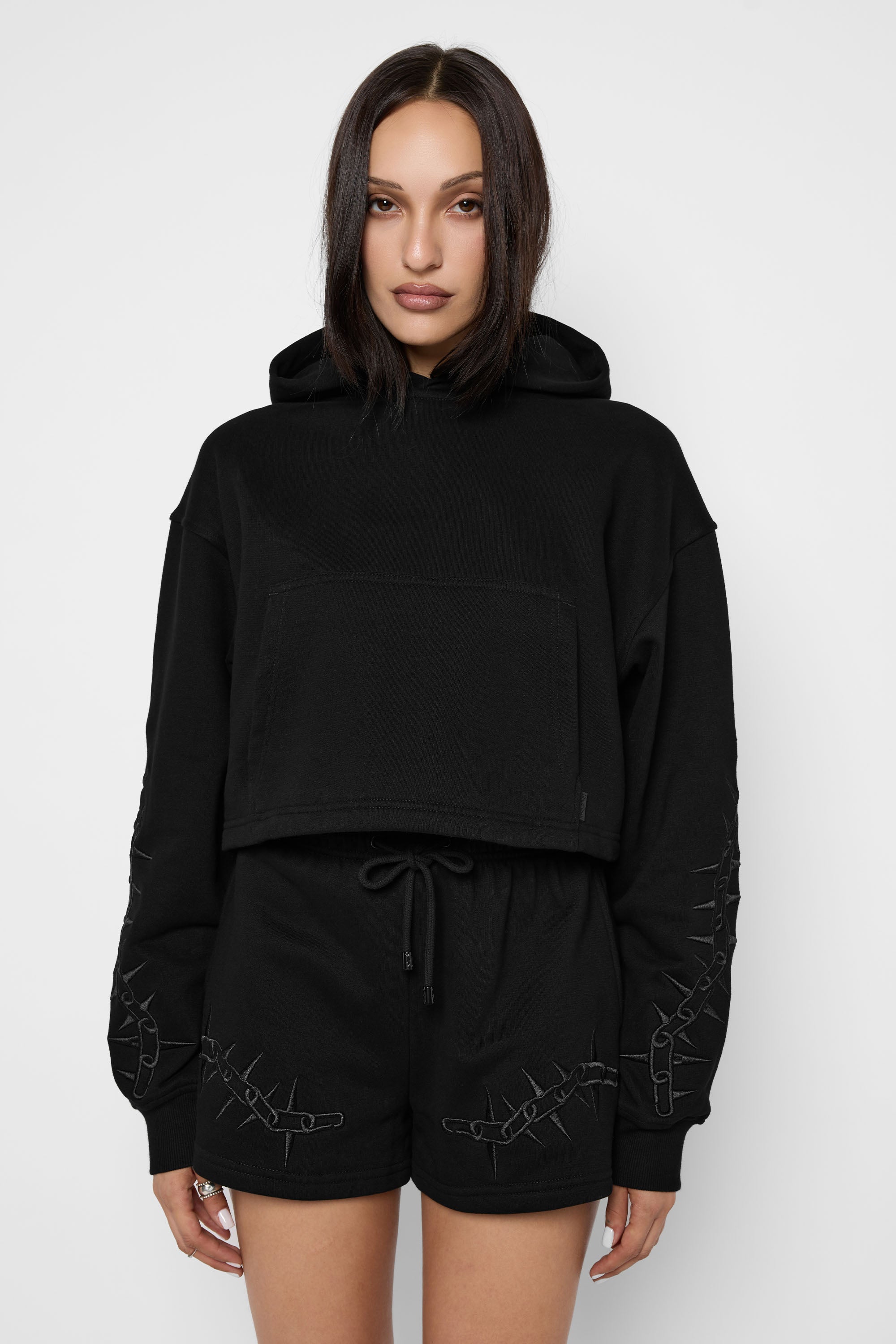 A model wearing a black cropped pullover hoodie with a half-zip front, a large front pouch pocket, and matching black shorts featuring a drawstring waist and distressed graphic details on the sleeves and hem, shot against a plain white studio background.